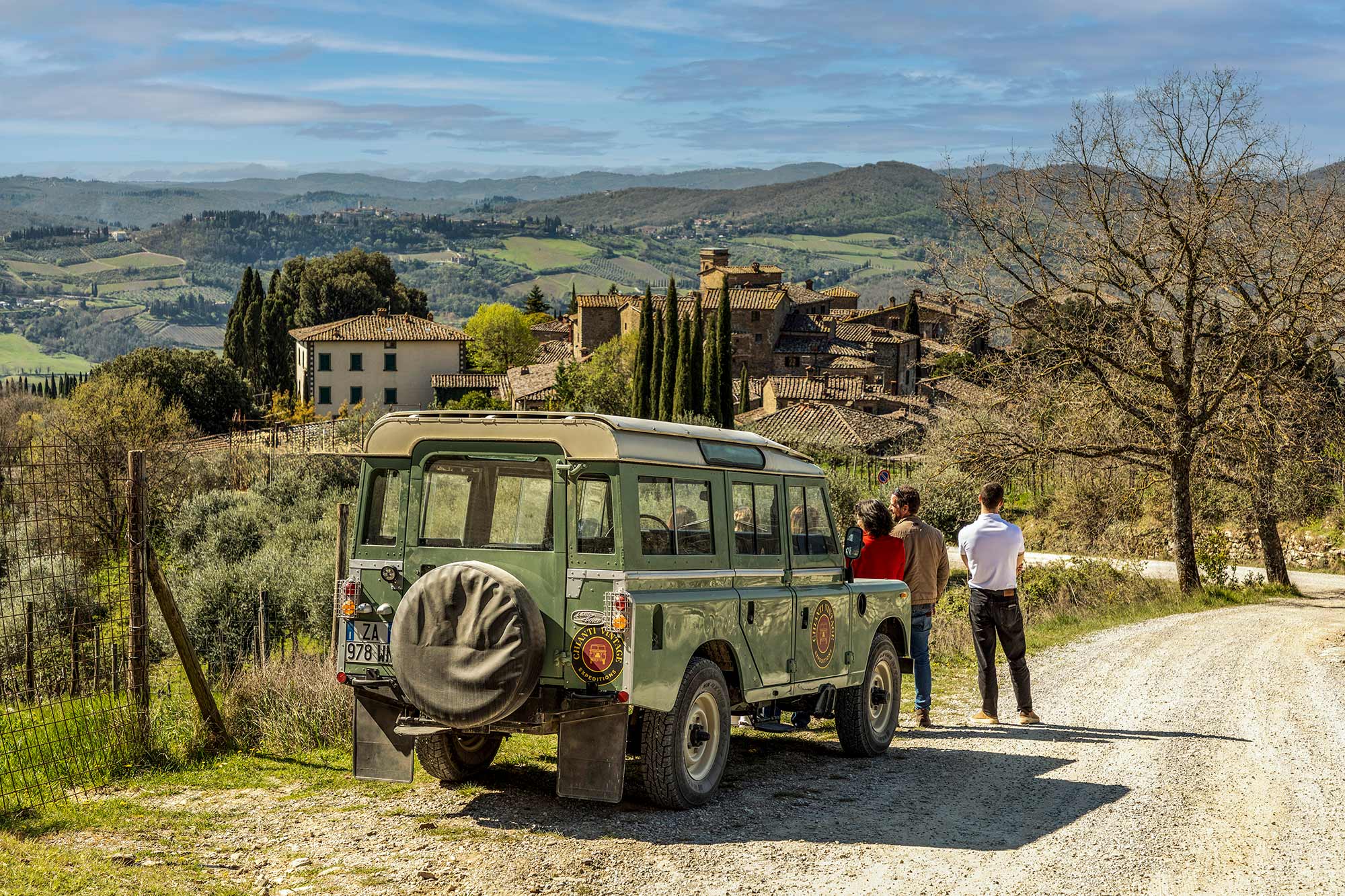 Due ospiti accanto alla Land Rover su una strada sterrata con un borgo medievale sullo sfondo