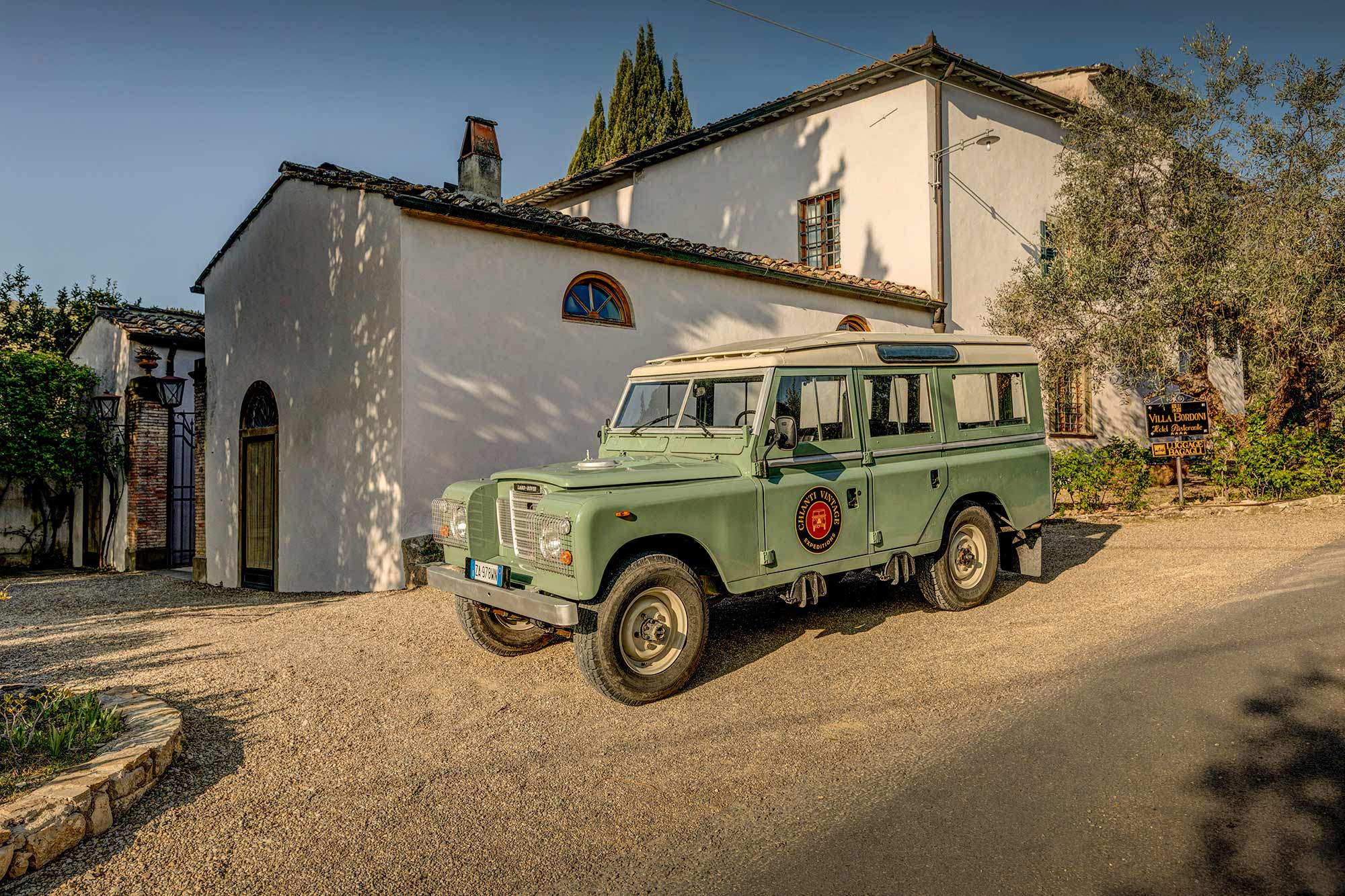 Land Rover Series III d'epoca davanti a una casa colonica toscana bianca al tramonto