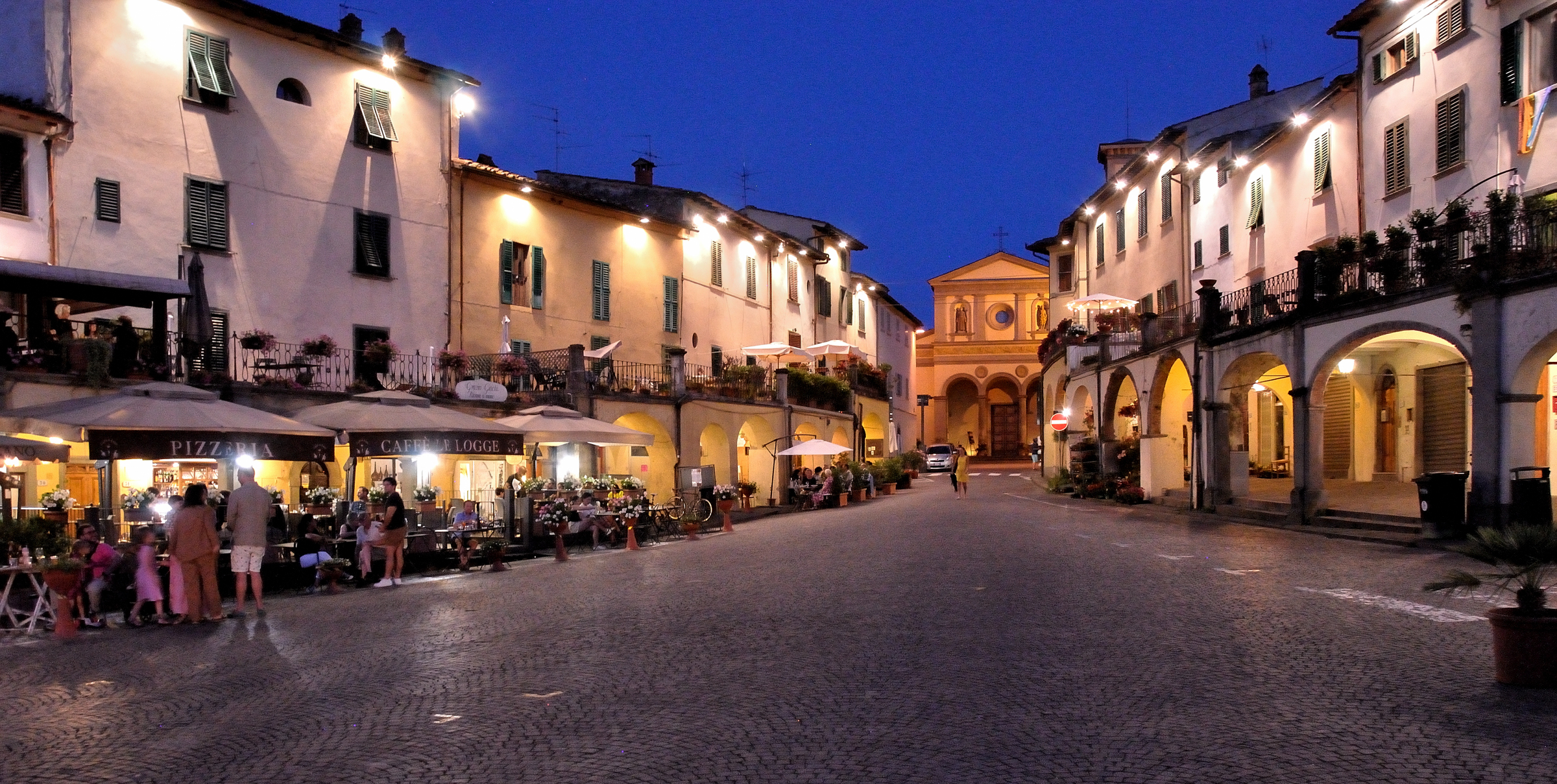 La piazza porticata di Greve in Chianti illuminata di sera con ristoranti e gente a passeggio