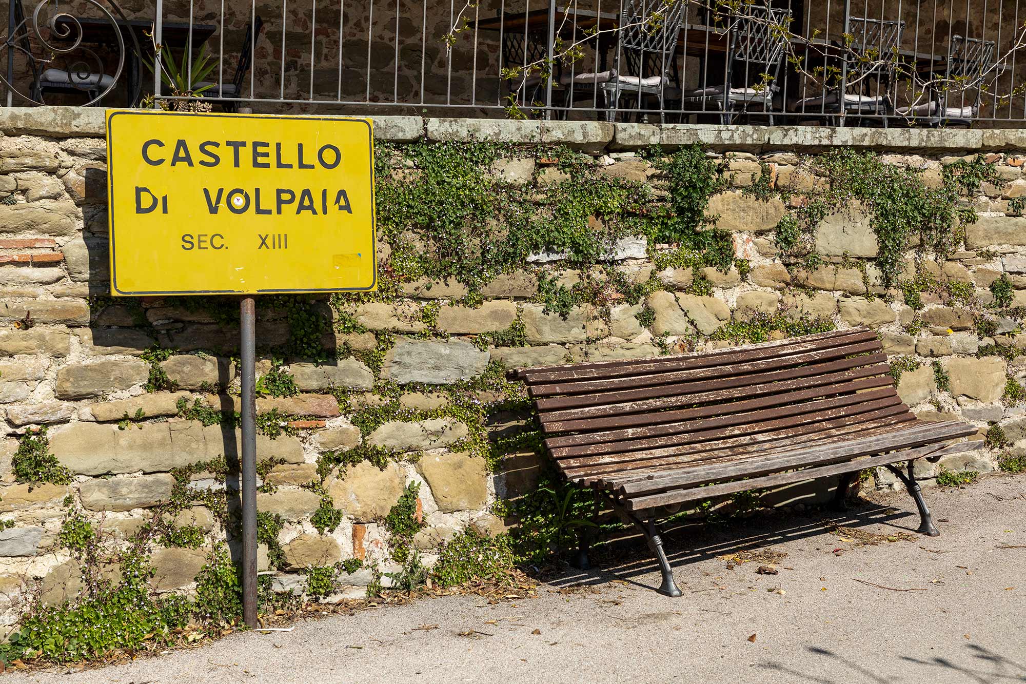 Cartello giallo Castello di Volpaia Sec. XIII su muro in pietra con edera e panchina di legno