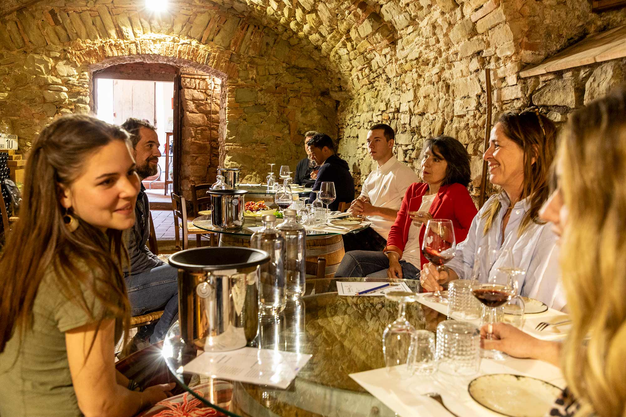 Gruppo di ospiti a cena attorno a un lungo tavolo in una cantina sotterranea a volta in pietra